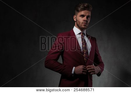 portrait of handsome man buttoning his red suit and looking to side while standing on grey wallpaper background