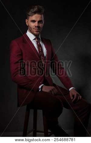 stylish man in grena suit sitting on a wooden chair and smiling on grey wallpaper background