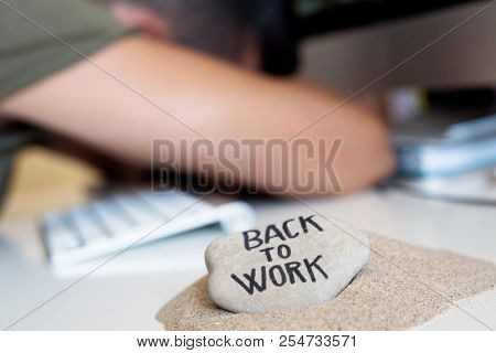 closeup of a concerned man sitting at his office desk, and stone on a pile of sand in the foreground with the text back to work written in it