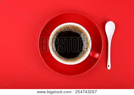 Close Up One Full Cup Of Black Americano Coffee And Saucer Ith White Spoon Over Vivid Red Paper Back