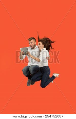 Jump Of Young Couple Over Red Studio Background Using Laptop Or Tablet Gadget While Jumping. Runnin 