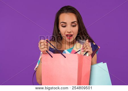 Beautiful Woman Peeks Into A Package, In The Hands Of Multi-colored Shopping Bags On A Purple Backgr