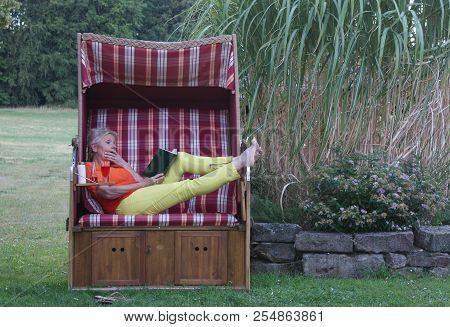 Joyfully With A Laugh On Her Face, The Blonde Woman Reads In The Humorous Book
