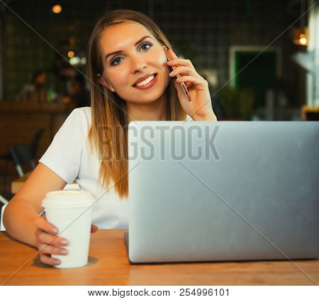 Young beautiful blond girl sitting in a coffee shop and working on computer. Image of happy woman using laptop and smartphone in cafe. Lifestyle concept.