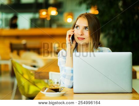 Young beautiful blond girl sitting in a coffee shop and working on computer. Image of happy woman using laptop and smartphone in cafe. Lifestyle concept.