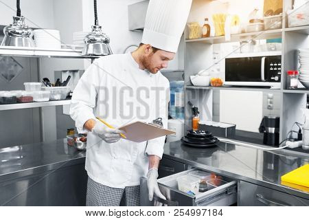 cooking, profession and people concept - male chef cook with clipboard doing inventory and looking to fridge at restaurant kitchen