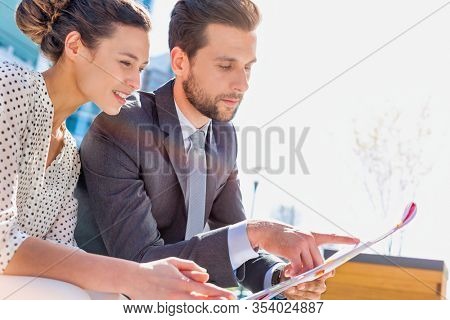 Young attractive businessman discussing business plans over document with beautiful businesswoman while sitting on bench against office building