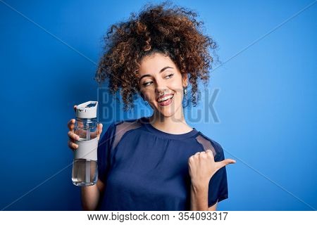 Young beautiful sporty woman with curly hair and piercing doing sport holding bottle of water pointing and showing with thumb up to the side with happy face smiling
