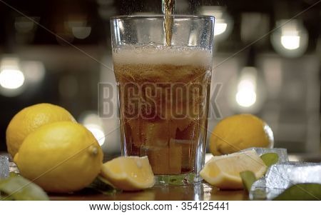 Close Up Fizzy Drink Pouring Into The Glass. Drink With Ice And Lemon In Glass On Blurry Lights Back