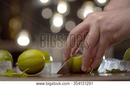 Close Up Male Hand Slicing Lime On The Table Over Background Of Blurry Light Bulbs. Cooking Of Refre