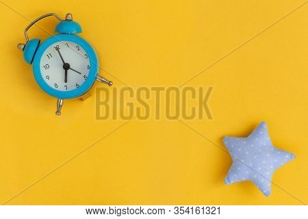 Soft Blue Pillow In Shape Of Star And Blue Alarm Clock On Yellow Background. Flat Lay.