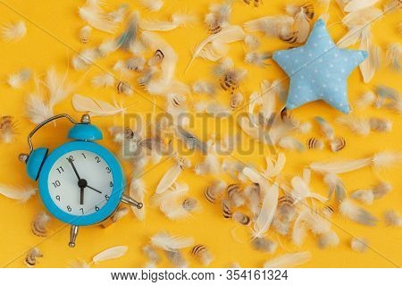 Soft Blue Pillow In Shape Of Star And Blue Alarm Clock And Feathers On Yellow Background. Flat Lay.