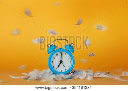 Soft Fluffy Flying Feathers And Blue Alarm Clock On Yellow Background. Wake Up Concept.