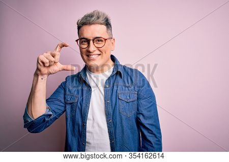 Young handsome modern man wearing glasses and denim jacket over pink isolated background smiling and confident gesturing with hand doing small size sign with fingers looking and the camera. Measure 