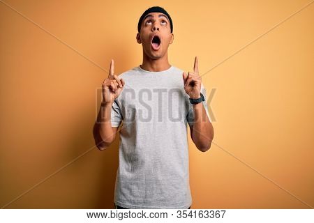 Handsome african american sportsman doing sport wearing sportswear over yellow background amazed and surprised looking up and pointing with fingers and raised arms.