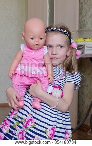 Caucasian Blond Girl With Hairstyle In A Striped Dress Plays With A Doll In Pink Clothes