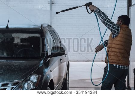 Young Man Washing Gray Car With Water Hose At Outdoors Car Wash Close Up. Wet Car In Water Droplets 