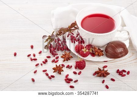 Healthy fruit tea with anise, barberries and raspberries in white porcelain cup and branch of berberry on light wooden background.