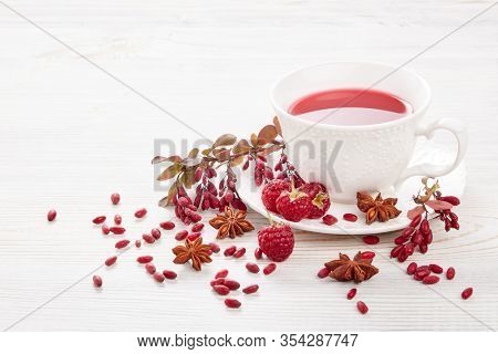 Healthy fruit tea with anise, barberries and raspberries in white porcelain cup and branch of berberry on light wooden background.