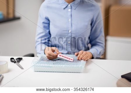 delivery, mail service, and shipment concept - close up of woman sticking fragile mark to parcel in protective bubble wrap at post office