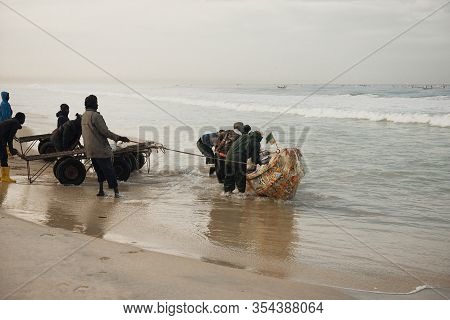 Kayar, Senegal - January 17, 2020: Fishermans Pushing Traditional Painted Wooden Fishing Boat In Kay