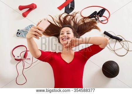 Top View Of Sporty Woman Laying On White Surface With Happy Facial Expression, Making Selfie Via Her
