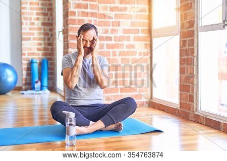 Middle age handsome sportman sitting on mat doing stretching yoga exercise at gym with hand on headache because stress. Suffering migraine.