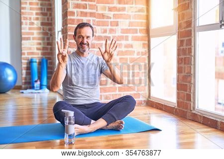 Middle age handsome sportman sitting on mat doing stretching yoga exercise at gym showing and pointing up with fingers number eight while smiling confident and happy.