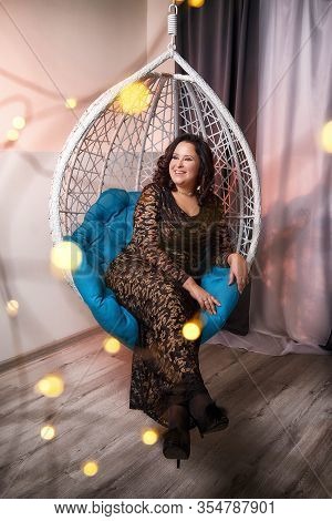 Fat Plump Charming Cute Woman With Black Curly Hair In Black Beautiful Dress In A White Wicker Chair