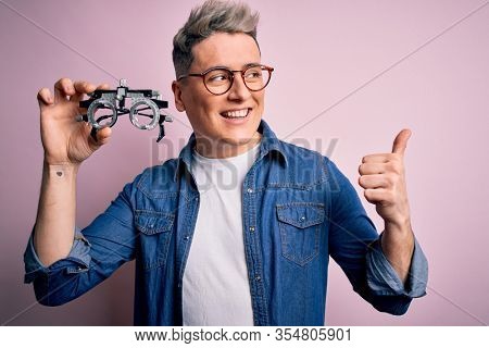 Young modern optical optomestrist man holding optometrical glasses over pink background pointing and showing with thumb up to the side with happy face smiling