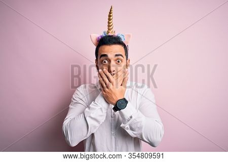 Young handsome man with beard wearing funny unicorn diadem over pink background shocked covering mouth with hands for mistake. Secret concept.