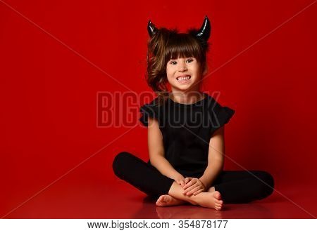 Little Brunette Girl With Devil Horns, Ponytail, In Black Blouse And Leggings, Barefoot. She Is Smil