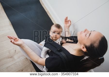 Young Mother Doing Yoga Exercises And Meditation With Her Little Baby In Ergo Backpack.