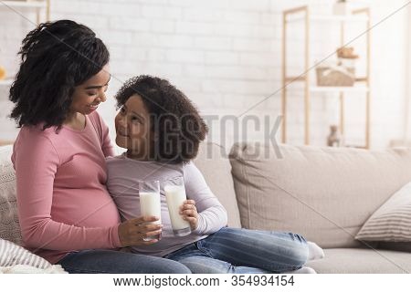 Little African American Girl And Her Pregnant Mother Drinking Milk Sitting On Sofa At Home, Clinking