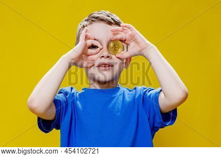 Little Happy Preschool Boy In A Blue T-shirt Holding Bitcoin In Front Of His Eyes On Yellow Backgrou