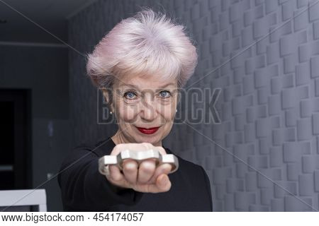 An Elderly Woman Strikes With Brass Knuckles On A Light Background, Close-up, Selective Focus. Conce