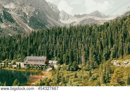 Wooden Cottage In The Mountains And Green Coniferous Forest Against The Backdrop Of Pale Gray Mounta