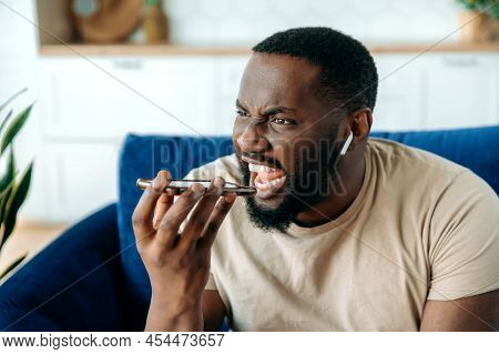 Outraged Angry Outraged African American Guy, Sitting On A Sofa In The Living Room, Uses Wireless Ea