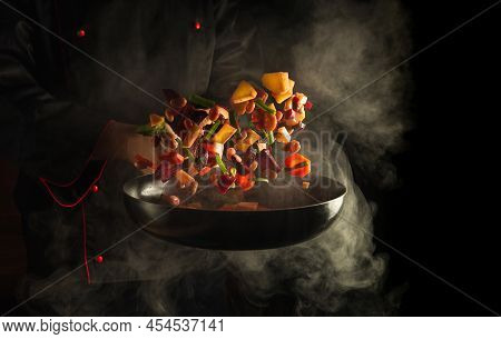 Professional Chef Prepares Food In A Hot Pan With Steam On A Black Background. The Concept Of Restau