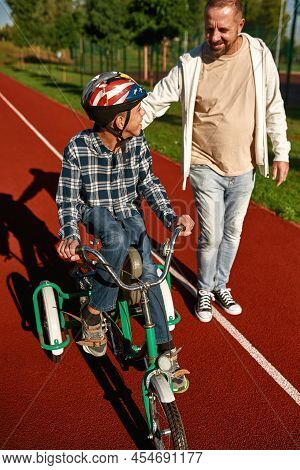 Caucasian Father Going And Helping Son With Cerebral Palsy Riding Tricycle In Sunny Park. Man And Bo