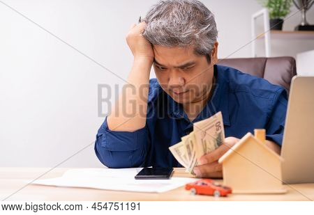 Unhappy Asian Man Holding A Banknote And Making An Account Of Income And Expenses To Pay The Home Lo