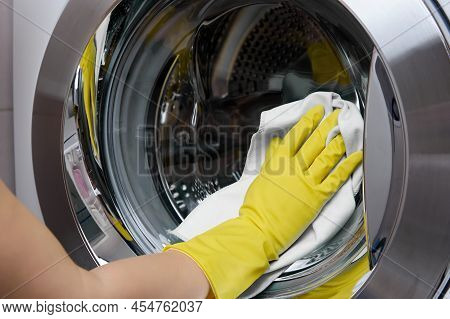 In The Bathroom, A Cleaner Wipes A Steel-colored Washing Machine Drum With A White Rag. A Womans Han