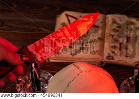 Close-up Hand Holds Knife Stained With Blood On The Satanic Book Background. Antichrist Satanic Ritu