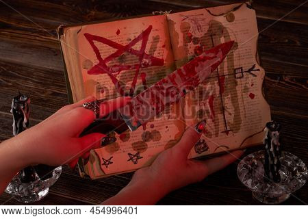 Satanic Wicca Ritual Concept. Female Hand Holds Bloody Knife Over The Bible With Pentagram.