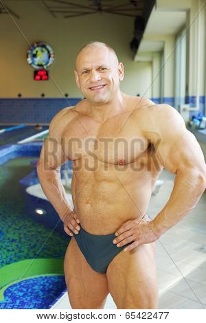 Happy bodybuilder in swimming trunks stands near indoor pool of gym hall