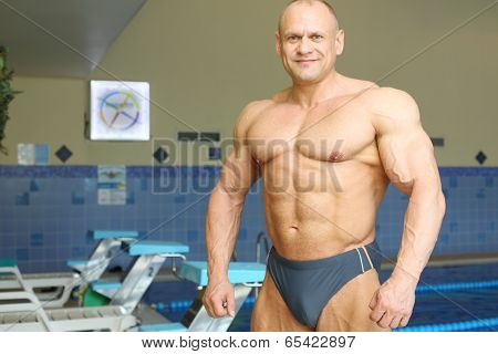 Smiling bodybuilder stands near indoor pure pool before swimming 