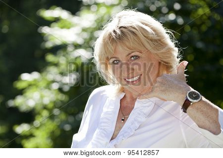 Happy smiling old blonde woman with call me gesture, against green sunner park