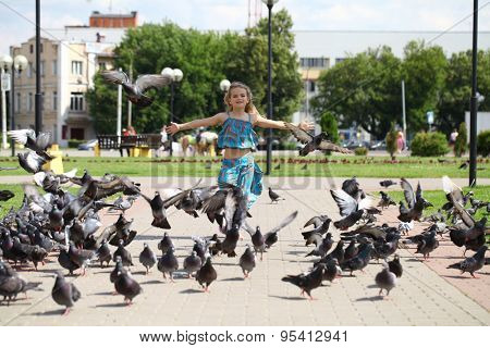 Young happy girl runs through a flock of pigeons on the square in summer city park