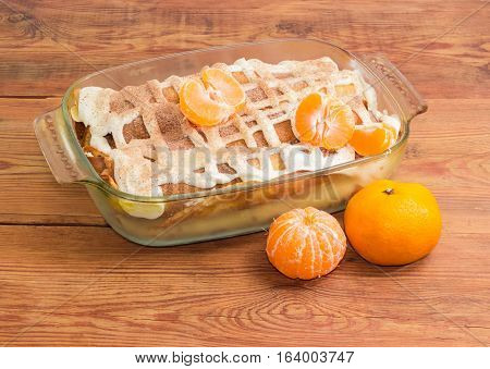 Sweet pancakes filled with cottage cheese with sour cream and sprinkled with cinnamon powder in the rectangular glass roasting pan and mandarins on old wooden surface
