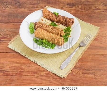 Pancakes filled with meat filling and parsley twigs on a white dish and fork on a cloth napkin on an old wooden surface